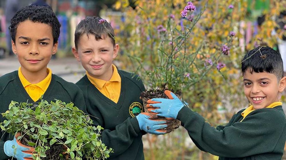 Three pupils hold up plants to the camera. They are dressed in green and  yellow school uniform
