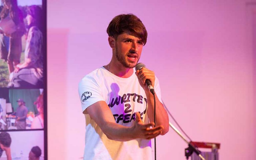 A man performs poetry wearing a t-shirt that says Write to Speak