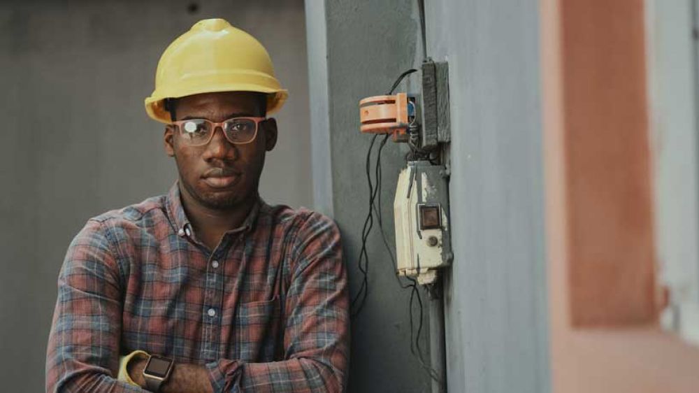 A young black man in glasses stares out of the camera. He is wearing a builder's hard hat.