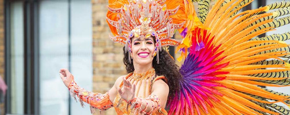 A carnival performer dressed in bright feathers dances and looks straight down the camera