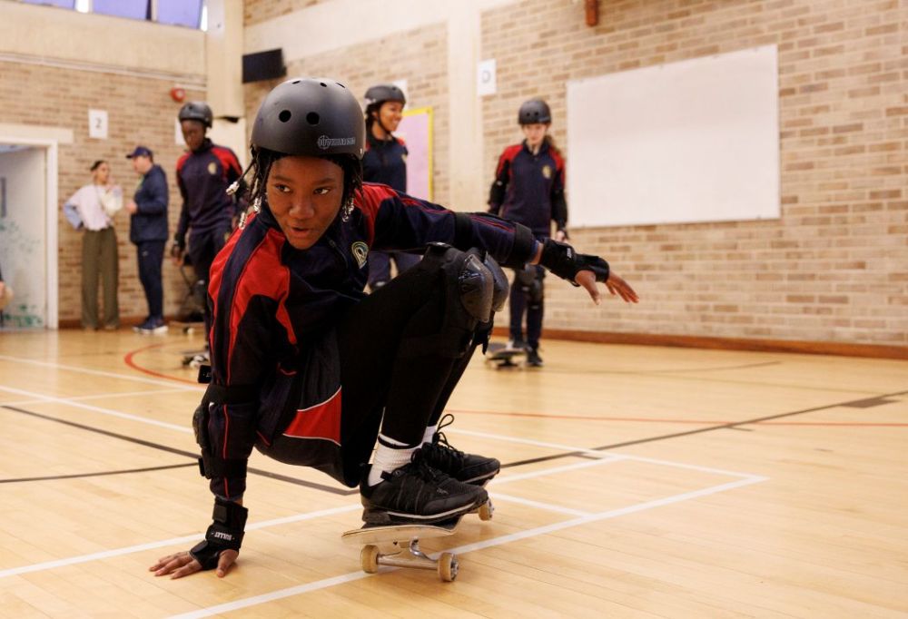 A girl in PE Kit and helmet skates low on a skateboard