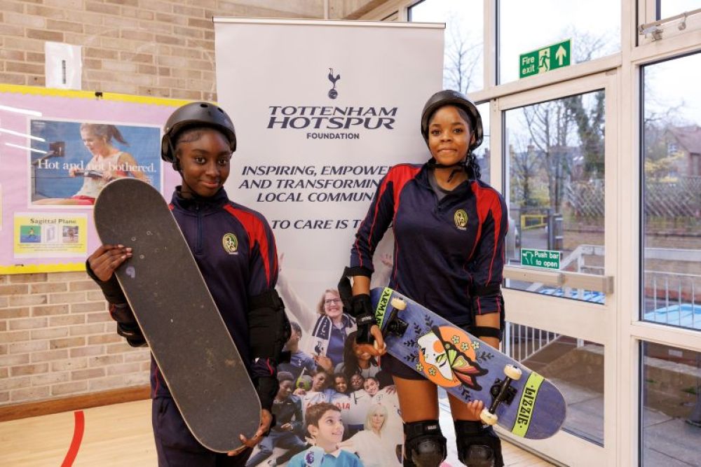 Two girls in PE kits hold up their skateboards. THFC banner is behind them. 