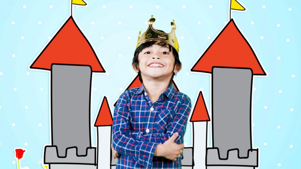 A boy stands in front of a drawing of a castle. He wears a paper crown. 