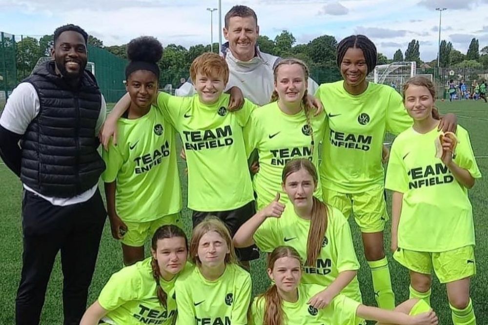 Girls football team pose with 2 adults. The team members are wearing bright yellow Team Enfield t-shirts