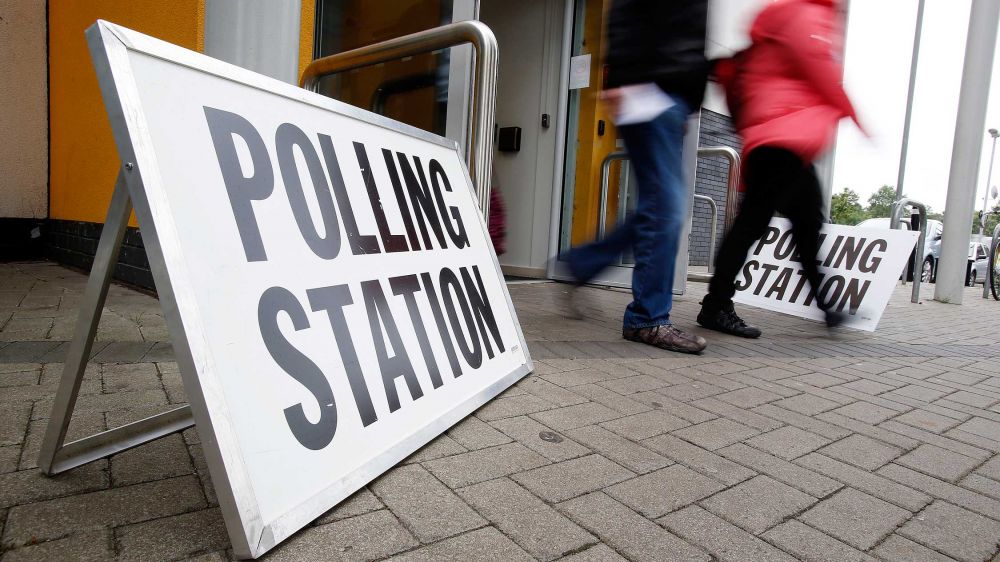 A sign for a polling station rests on the ground against an open door to a building. Voters are exiting the building in the background.